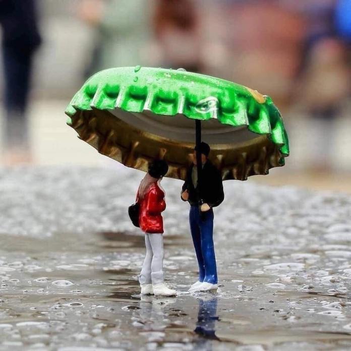 Miniature figures of a man and woman standing beneath a green bottle cap used as an umbrella, placed on wet pavement with visible water droplets, in a rainy city street scene.