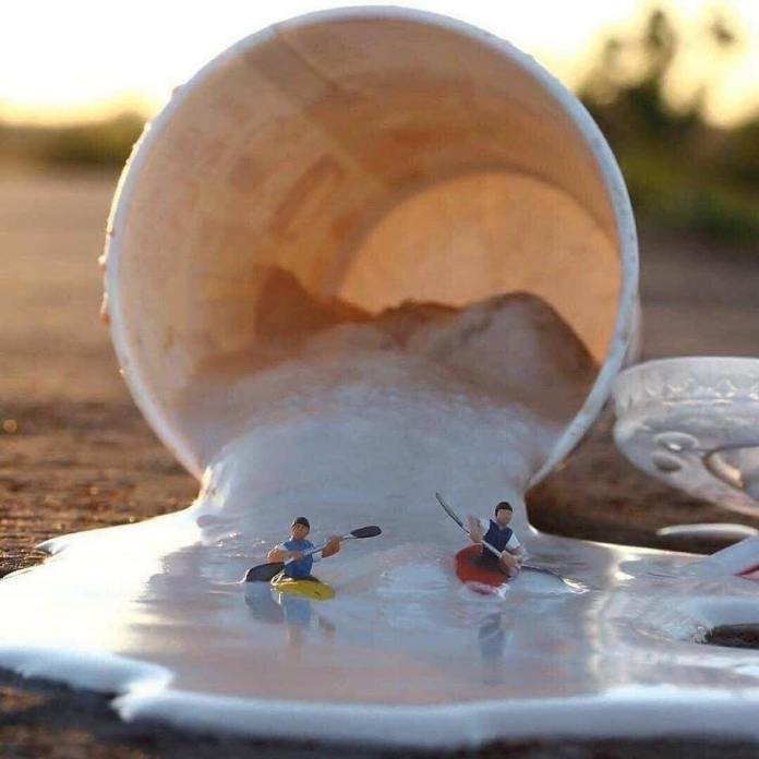Two miniature kayakers paddling on a white liquid flowing from a tipped-over cup, simulating rapids in a playful urban river scene.