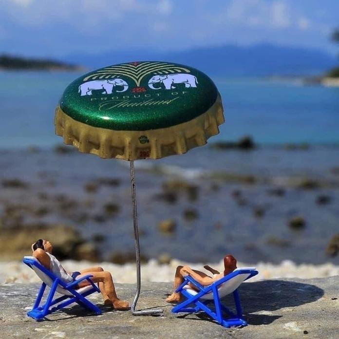 Miniature figures reclining on blue beach chairs under a green Thai bottle cap used as a beach umbrella, with the ocean and distant mountains behind them.