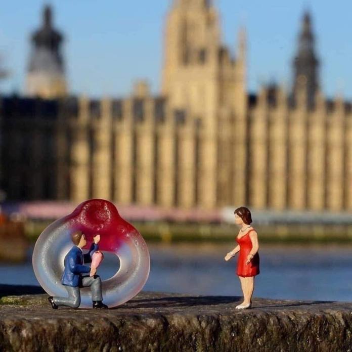 Miniature man kneeling with a large red-and-clear candy ring proposing to a woman in a red dress, with the UK Parliament buildings in soft focus behind.
