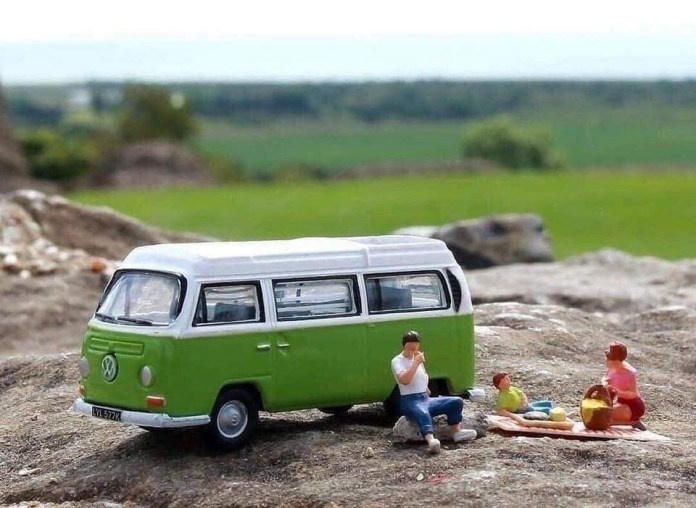 Tiny family figures picnicking next to a green VW camper van toy, with food on a blanket and natural scenery in the background.