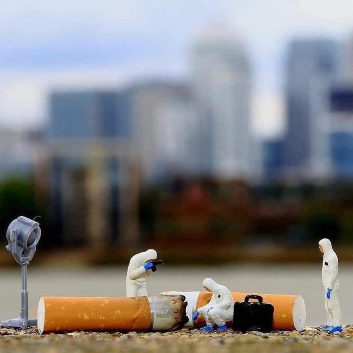 Miniature hazmat-suited figures investigating a large cigarette butt as a mock crime scene, with city buildings blurred in the background.