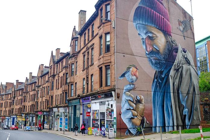 Street mural by the artist SMUG on a building in Glasgow, Scotland, depicting an elderly man with a gentle expression, wearing a red knit beanie and a worn jacket. He holds a small robin on his finger, which gazes back at him, creating a touching connection between man and nature. Another robin hovers nearby, adding to the scene's warmth and intimacy. The artwork seamlessly integrates with the building's architecture, bringing life and character to the street.