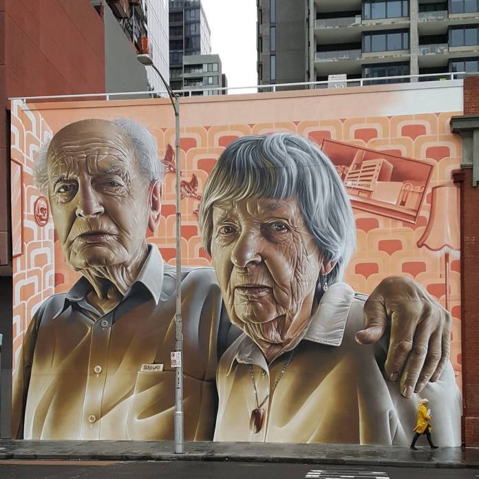 Detailed mural by SMUG in Melbourne, Australia, honoring the artist’s grandparents. The artwork features realistic, large-scale portraits of an elderly man and woman, both looking directly at the viewer with expressions that capture wisdom and resilience. The man’s hand rests gently on the woman’s shoulder, symbolizing connection and support. Set against a warm, patterned background that includes nostalgic elements, the mural celebrates family and heritage in SMUG’s hyper-realistic style.
