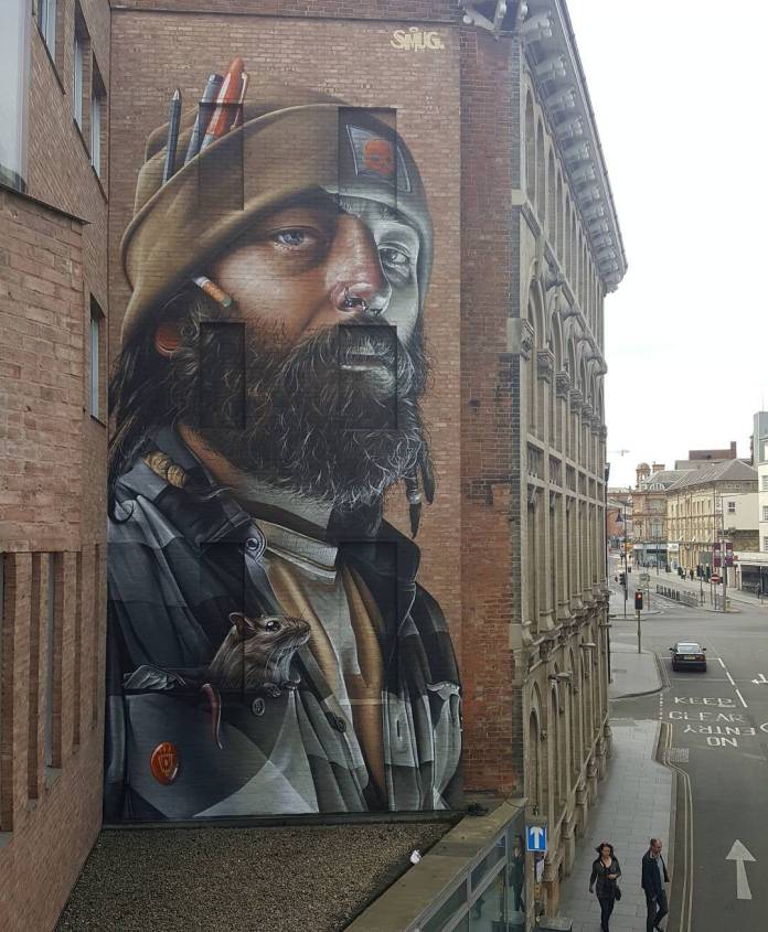 Large mural by SMUG on the side of a building in Leicester, UK, created for the Bring The Paint Street Art Festival. The artwork depicts a bearded man wearing a beanie with pencils and a paintbrush tucked into it. A small rat peeks out from his jacket pocket, adding a whimsical touch. The detailed realism in SMUG’s style brings depth and character to the portrait, blending hyper-realistic features with subtle storytelling.