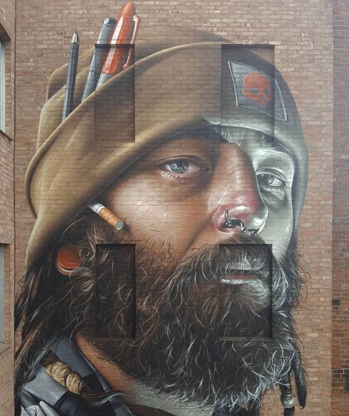 Close-up view of a large mural by SMUG at the Bring The Paint Street Art Festival in Leicester, UK. The artwork features a bearded man wearing a beanie adorned with pencils, a paintbrush, and a cigarette tucked behind his ear. The realistic detail in the texture of the beard, skin, and fabric showcases SMUG's impressive photorealistic style. The mural uses the building's natural architectural lines to enhance the depth and expression in the man's face.
