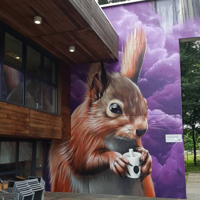 A mural of a squirrel holding a coffee cup with an inquisitive expression, painted against a purple cloud backdrop. The squirrel’s fur appears incredibly lifelike, with intricate brushstrokes capturing its texture. This artwork adds a cozy, endearing touch to the urban setting, created for a Street Art Festival in Belgium.