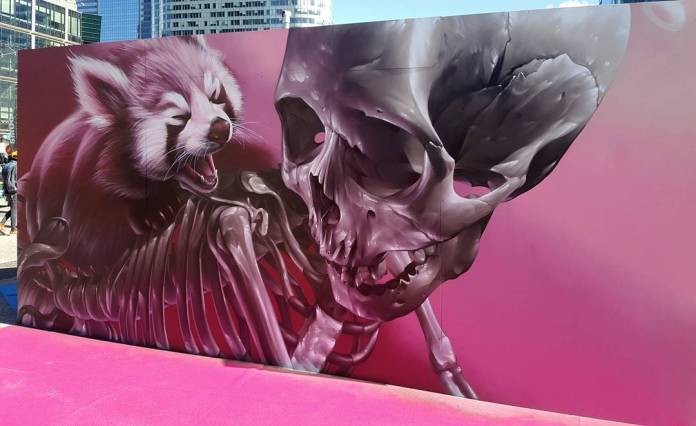 Mural by artist SMUG featuring a highly detailed, surreal image of a red panda with an exposed skeletal structure, roaring next to a large, intricately painted human skull. The artwork is set against a vibrant pink background, creating a striking contrast. Created for Projet SAATO in Paris, France, this piece combines elements of life and death in a captivating, otherworldly style.