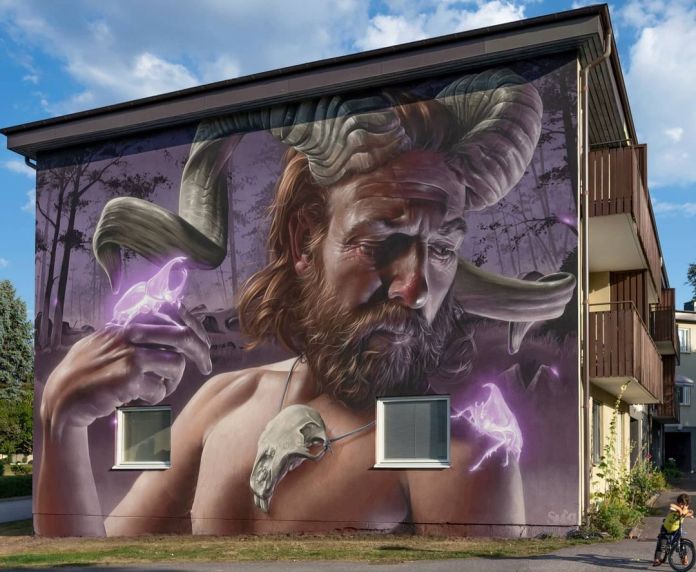 A mural by SMUG located in Västervik, Sweden at BANK! Västervik Street Art, depicting a bearded man with a contemplative expression, wearing large ram horns and holding his hand up to display a glowing purple figure of a small animal. The man’s necklace also holds a skull, adding a mystical and mythical feel to the artwork. The background features a dark forest, blending realism with fantasy elements, creating an ethereal atmosphere.