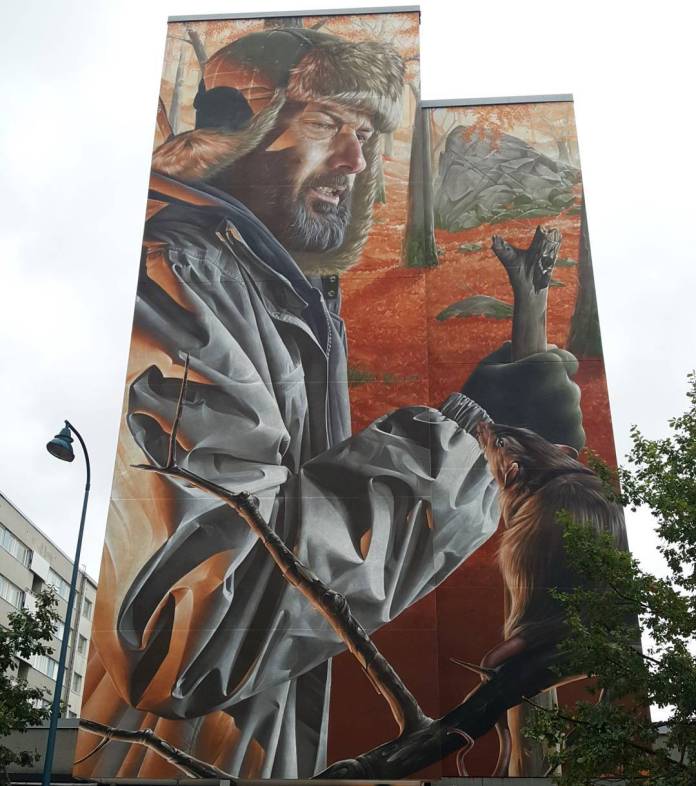 A mural by SMUG at Upeart in Kotka, Finland, depicting a rugged bearded man dressed in a heavy winter jacket with a fur-lined hood, holding a branch while a curious rat climbs toward his gloved hand. The background is a vibrant autumn forest scene with red and orange leaves on the ground, enhancing the realistic detail of the man's clothing and the natural environment. The artwork merges elements of nature and human connection in a lifelike yet whimsical portrayal.