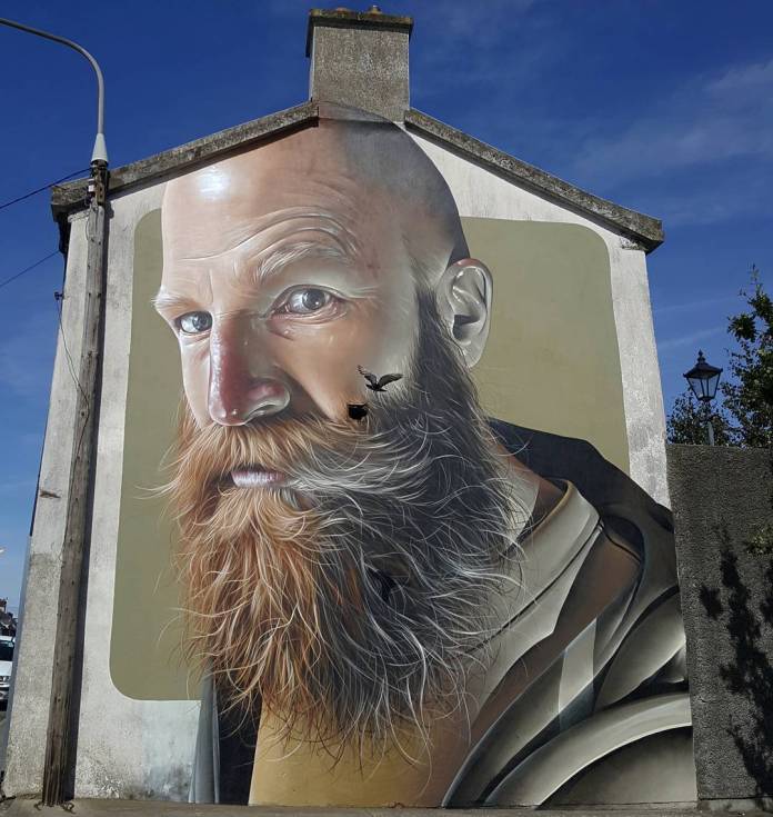 A mural by SMUG at the Waterford Walls Street Art Festival in Ireland, showcasing an ultra-realistic portrait of a bald man with a full, thick ginger beard and piercing blue eyes. The man’s expression is intense and contemplative, with lifelike detailing in his facial features and beard. A small bird appears to interact with the portrait, as though flying out from the man’s beard, adding a whimsical touch to the hyper-realistic artwork. The mural is painted against a muted background on the side of a building, with a streetlamp and a clear blue sky visible nearby.