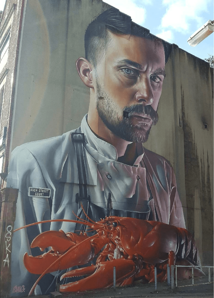 A hyper-realistic mural by SMUG in Belfast, Northern Ireland, depicting a close-up portrait of a chef with a contemplative expression, clad in a white chef's jacket with suspenders. In front of him is an oversized, vibrant red lobster, adding a surreal touch to the artwork. The mural showcases SMUG’s signature attention to detail, with intricate textures in both the chef’s facial features and the lobster's shell. The artwork is set on a tall building wall near a street corner, marked by a 'High Street Court' sign, with a cloudy sky visible above.