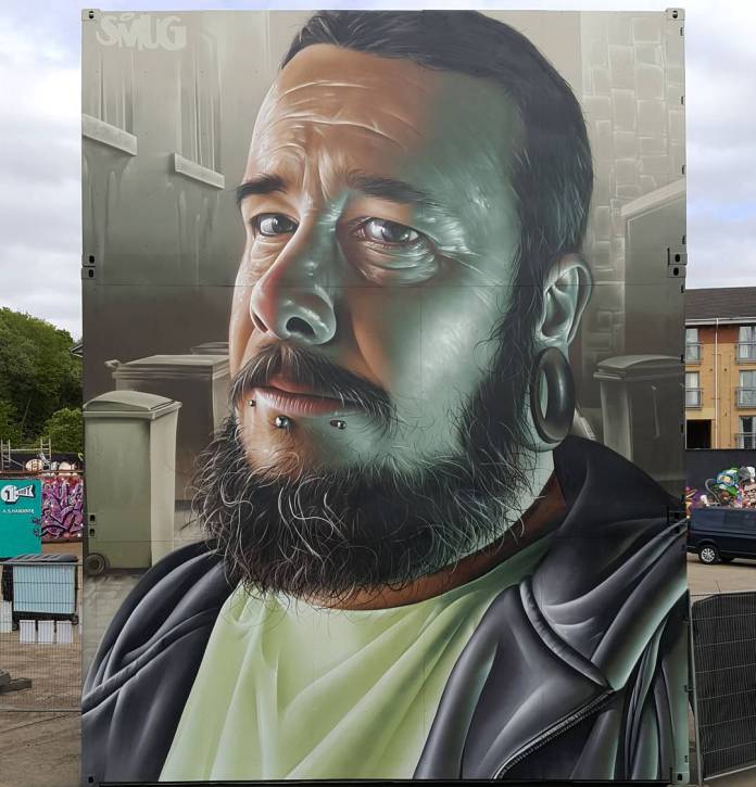 A striking mural by SMUG at Yardworks in Glasgow, Scotland, portraying a hyper-realistic close-up of a man with a beard, piercings, and stretched earlobe piercings. The subject's expression is thoughtful, with subtle shadows and highlights enhancing the texture of his skin and beard. He wears a dark jacket over a light shirt, and the background features a muted, urban setting with soft lighting. SMUG's attention to detail is evident in the lifelike textures and nuanced shading that give the mural a three-dimensional feel.