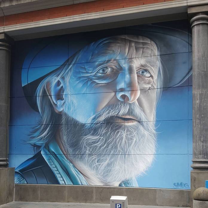Mural by SMUG in Melbourne, Australia, featuring a close-up portrait of an older man with a long, textured white beard and contemplative blue eyes. The artwork uses cool tones, primarily blue and white, giving the subject an ethereal, almost serene look. The man wears a hat and a jacket, with detailed shading and highlights enhancing the realism of his weathered skin and beard. The mural is set between two stone pillars, adding depth to the artwork. SMUG's precision in capturing lifelike textures and expressions is evident in this striking portrait.