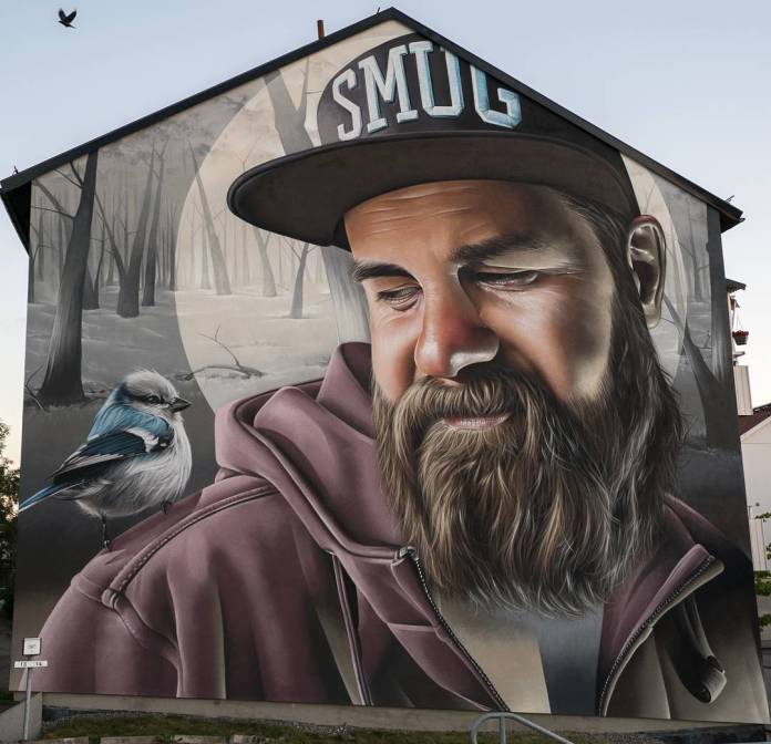 Large mural by SMUG for Artscape in Årjäng, Sweden, featuring a close-up portrait of a bearded man wearing a hoodie and a cap with 'SMUG' written on it. The man looks down softly at a small blue and white bird perched on his shoulder, creating a sense of peaceful interaction. The background shows a misty, monochromatic forest, enhancing the serene and introspective mood of the mural. The artist's attention to detail is evident in the realistic textures of the man’s beard, skin, and clothing, as well as the soft feathers of the bird.