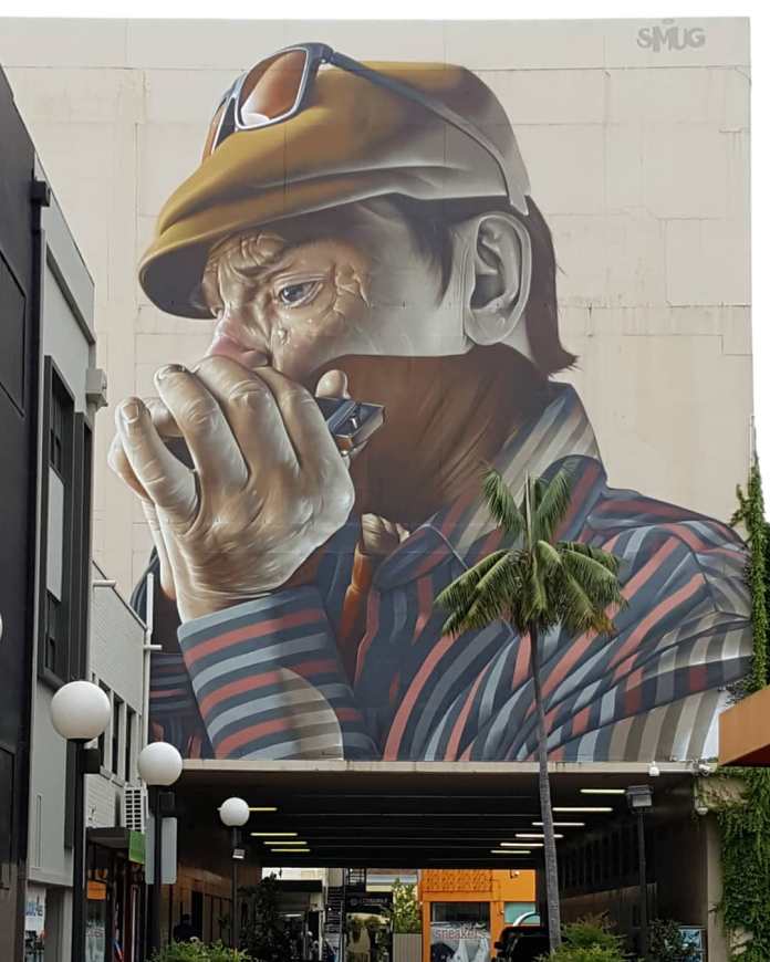 Mural by SMUG at the Wonderwalls Festival in Wollongong, Australia, depicting a close-up of a man playing a harmonica. The man wears a cap with sunglasses resting on top and a striped shirt, his face deeply focused on the music. The intricate details capture his facial wrinkles, expression, and the texture of his hands gripping the harmonica. This artwork, covering a large wall, showcases SMUG’s realistic style, with striking shading and color contrasts that bring the figure to life against the urban backdrop.