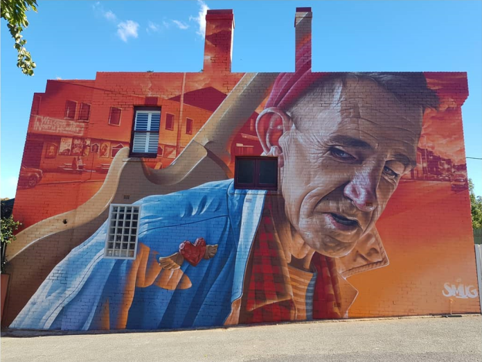 A detailed mural by Smug in North Hobart, Tasmania, Australia, depicting an elderly man named Cam with a deeply expressive face, set against a warm orange and red backdrop of the local neighborhood. The man has a well-defined, lined face and wears a blue shirt, with a guitar slung over his shoulder. A carved heart emblem is displayed on his chest, symbolizing his custom-made heart brooches crafted for local women. Smug's use of a vibrant, atypical color palette creates an inviting and nostalgic atmosphere that reflects the character and charm of Cam.