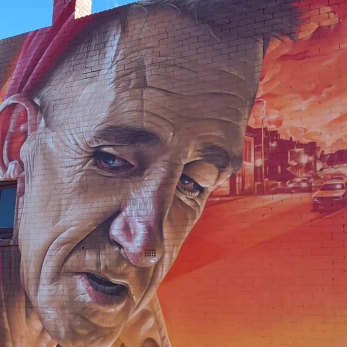 A detailed mural by Smug in North Hobart, Tasmania, Australia, depicting an elderly man named Cam with a deeply expressive face, set against a warm orange and red backdrop of the local neighborhood. The man has a well-defined, lined face and wears a blue shirt, with a guitar slung over his shoulder. A carved heart emblem is displayed on his chest, symbolizing his custom-made heart brooches crafted for local women. Smug's use of a vibrant, atypical color palette creates an inviting and nostalgic atmosphere that reflects the character and charm of Cam.