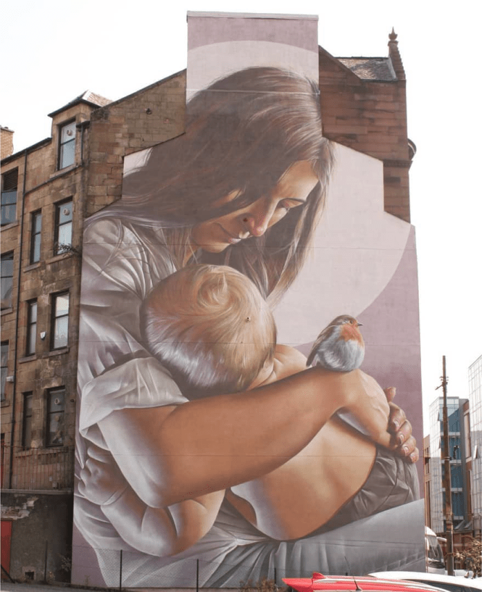 A touching mural by Smug located in Glasgow, Scotland, depicting a serene scene of a mother lovingly holding her young child. The mother’s face is gently inclined towards the child, embodying warmth and protection, while a small robin perches on her arm, adding a tender, peaceful element to the composition. Smug’s hyper-realistic style captures intricate details, such as the strands of hair and soft folds in the clothing, creating a lifelike portrayal against the textured wall of the building.