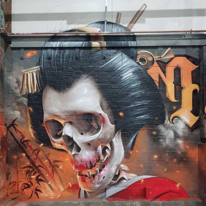 A striking mural by Smug located in Melbourne, Australia, featuring a skeletal figure with traditional Japanese Geisha elements. The figure's skull is adorned with a stylized black wig, chopsticks, and a small golden hairpiece, creating a unique blend of cultural references and macabre imagery. The background includes bamboo stalks and stylized orange and yellow hues, adding a fiery ambiance to the scene. Smug's detailed, hyper-realistic technique enhances the haunting yet captivating expression, with glowing embers floating around the skull, giving the mural a dynamic, intense feel.