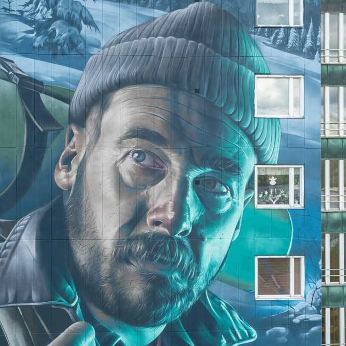 Close-up view of Smug's mural in Partille, Göteborg, for the Artscape project, inspired by the Finnish folk tale of Revontulet. The detailed portrait shows a bearded man wearing a knit beanie, with an intense expression as he looks off to the side, symbolizing the lost hiker from the story. The cool hues of blue and green enhance the icy, wintry setting, with snow-covered trees in the background. This mural, depicting the origin of the Northern Lights, is the tallest in Sweden and possibly the tallest in Scandinavia.