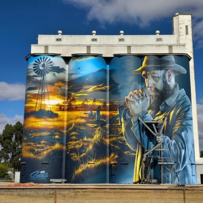 Mural on the Lameroo silos in South Australia, painted by street artist Smug. The artwork, curated by Juddy Roller, serves as a tribute to farmers, capturing the resilience and spirit of the Mallee farming community. The mural depicts a rugged farmer with hands clasped in contemplation, set against a vivid rural landscape at sunrise. A windmill and livestock are visible on the horizon, symbolizing the legacy and dedication of pioneering farmers in Lameroo. The warm golden tones blend with cool blues, creating a powerful contrast and emphasizing the hope and perseverance rooted in the community’s heritage.