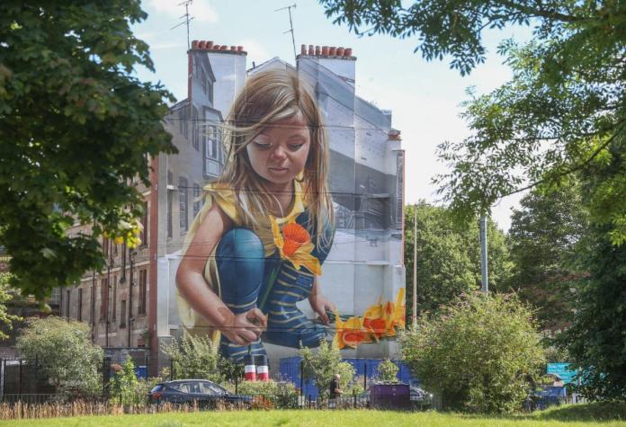 Mural by artist SMUG on a building in Glasgow, Scotland, created in collaboration with the Govan Housing Association to commemorate its 50th anniversary. The artwork depicts a young girl in overalls holding bright yellow daffodils, symbolizing the legacy of Peter Barr, the ‘Daffodil King’ from Govan. Barr is celebrated for his work in reviving the popularity of daffodils in the late 1800s, and a Memorial Cup is awarded annually to honor his contributions. The mural blends beautifully into the surrounding buildings, with greenery framing the scene.