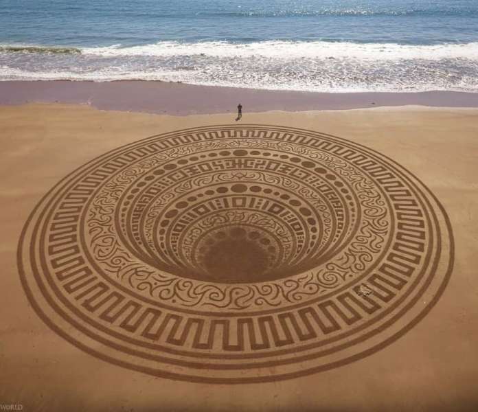 A breathtaking land art piece titled “Below” created by Jon Foreman at Lindsway Bay, Wales, UK. The design features an intricate circular pattern etched into the sand, with layers of geometric and organic motifs spiraling inward, creating the illusion of depth. A person stands near the edge, highlighting the massive scale of the artwork, with the ocean waves gently rolling in the background.