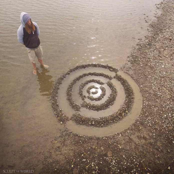 A land art piece titled “Extimus Lux” by Jon Foreman, featuring a spiral pattern created from stones placed in shallow water. The design begins at the center and radiates outward, forming a hypnotic swirl that blends with the natural shoreline. A person stands barefoot nearby, admiring the artwork, with the calm water reflecting light around the spiral.