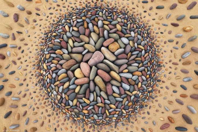 A vibrant land art piece titled “Explosia” by Jon Foreman, created at Freshwater West, Wales, UK. The design features a dense cluster of colorful stones arranged in a circular shape, radiating outward in smaller, scattered patterns. The intricate use of pebbles creates a sense of movement and energy, resembling an explosion captured in a still moment on the sandy beach.