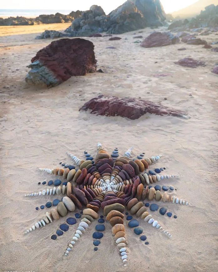 A radiant land art piece titled “Calefacio Stella” by Jon Foreman, created at Freshwater West, Wales, UK. The design resembles a starburst, crafted from carefully arranged stones of varying sizes and colors. The stones radiate outward in symmetrical, pointed formations, with the warm hues of the beach and distant rocks adding depth to the serene coastal setting.