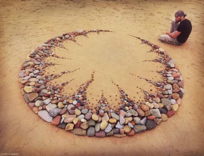 A captivating land art piece titled “Consumo” by Jon Foreman, created at Freshwater West, Wales, UK. The design forms a large circular arrangement of stones, with jagged, flame-like patterns extending inward toward the center. The stones vary in size and color, adding depth and texture to the piece. The artist sits nearby, reflecting on his work, blending perfectly with the serene beach setting.