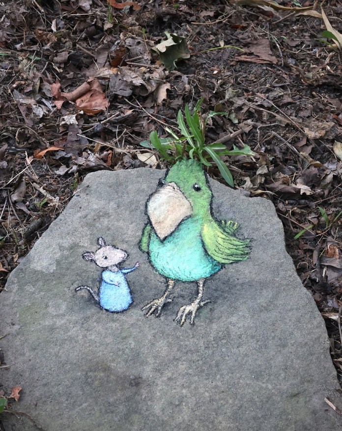 Chalk art of a small mouse in a blue sweater standing next to a large green bird drawn on a flat stone. A real clump of grass appears to sprout from the bird’s head like feathers.