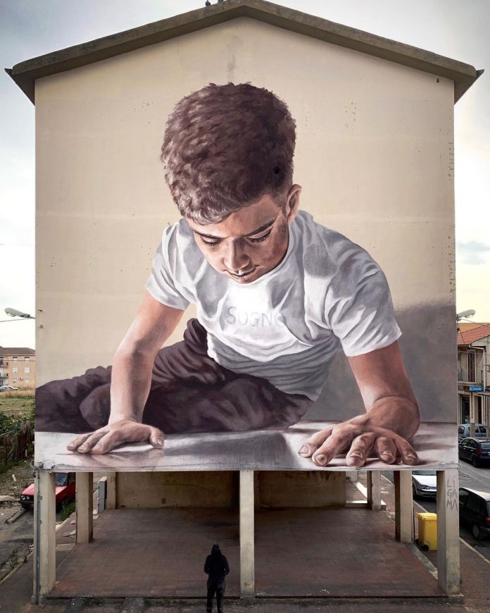 Photorealistic mural by LIGAMA in Ravanusa, Italy, depicting a large boy wearing a white t-shirt with the word "SOGNO" as he leans over a reflective surface that blends into the pavement, creating a 3D illusion.