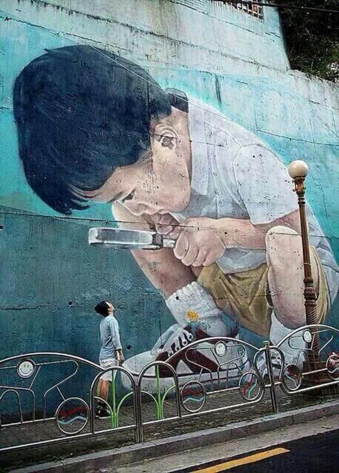 Large mural in Seoul, South Korea showing a young boy using a magnifying glass, painted with realistic detail on a blue wall, appearing to examine pedestrians walking below.