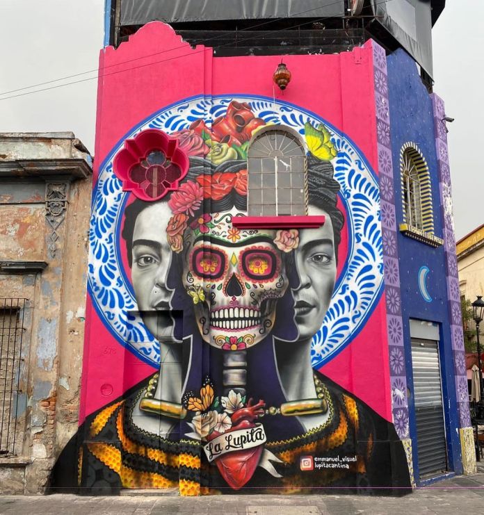 Mural on pink and purple building facade in Guadalajara with a stylized Frida Kahlo triple portrait, central skull in vibrant Día de los Muertos style, surrounded by flowers and Mexican folk patterns.