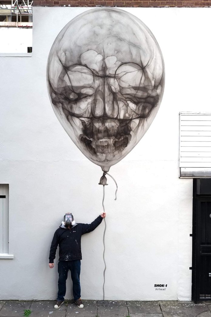 Mural of a translucent balloon shaped like a human skull, floating above a man holding its string. Spray-painted by SHOK-1 on a white wall in London using x-ray aesthetic.