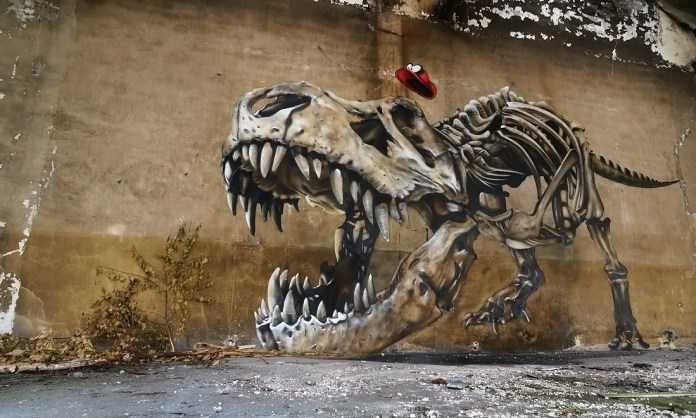 Street art of a large dinosaur skeleton painted with photorealistic shadows on a worn wall in Lorraine, France. A red light fixture is cleverly used as the creature’s eye.
