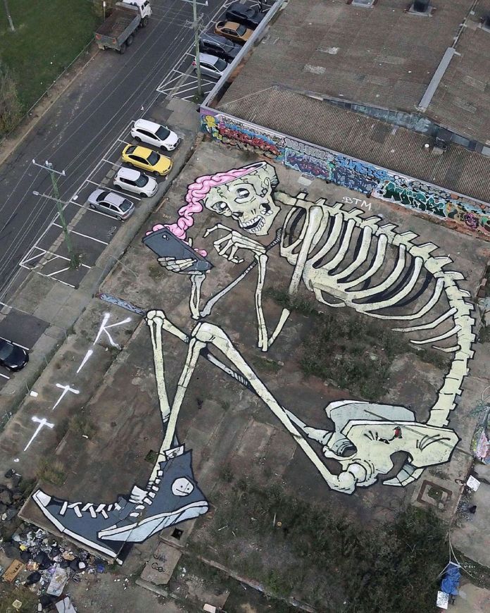 Gigantic skeleton mural painted on a vacant lot in Melbourne, Australia, with pink hair and sneakers, holding a smartphone in selfie pose. Created by Kitt Bennett.
