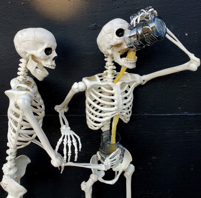Skeletons drinking beer (International Beer Day street art by ItSkeletal) – Two skeletons pose against a black wall; one pours beer into its skull, but the liquid spills through its ribcage into a mug held below. A humorous take on drinking without a stomach, created by street artist ItSkeletal.