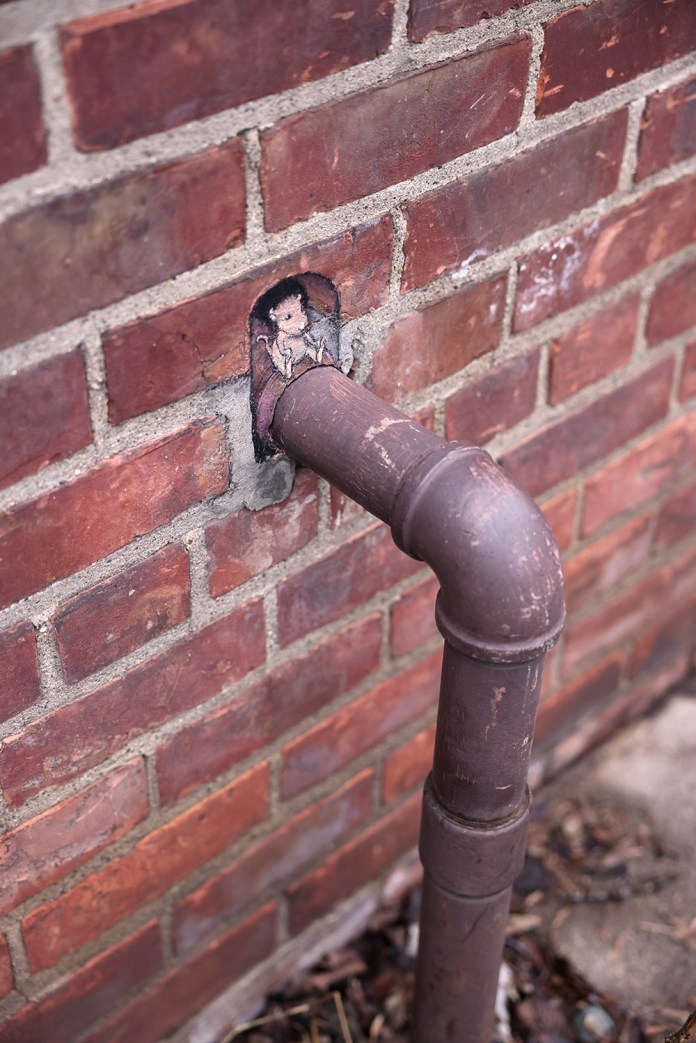 A charming illustration by David Zinn titled "Evelyn guards the pipe that sends kindness into the world." The artwork depicts a small, whimsical character nestled inside the opening of a pipe that extends from a red brick wall. Evelyn, with a gentle expression, appears to protect the pipe, blending seamlessly with the surrounding urban elements. The pipe, painted in earthy brown tones, complements the textured brick wall, creating a delightful interaction between the art and its environment. Bits of debris and foliage at the base of the pipe add a touch of natural contrast to the setting.