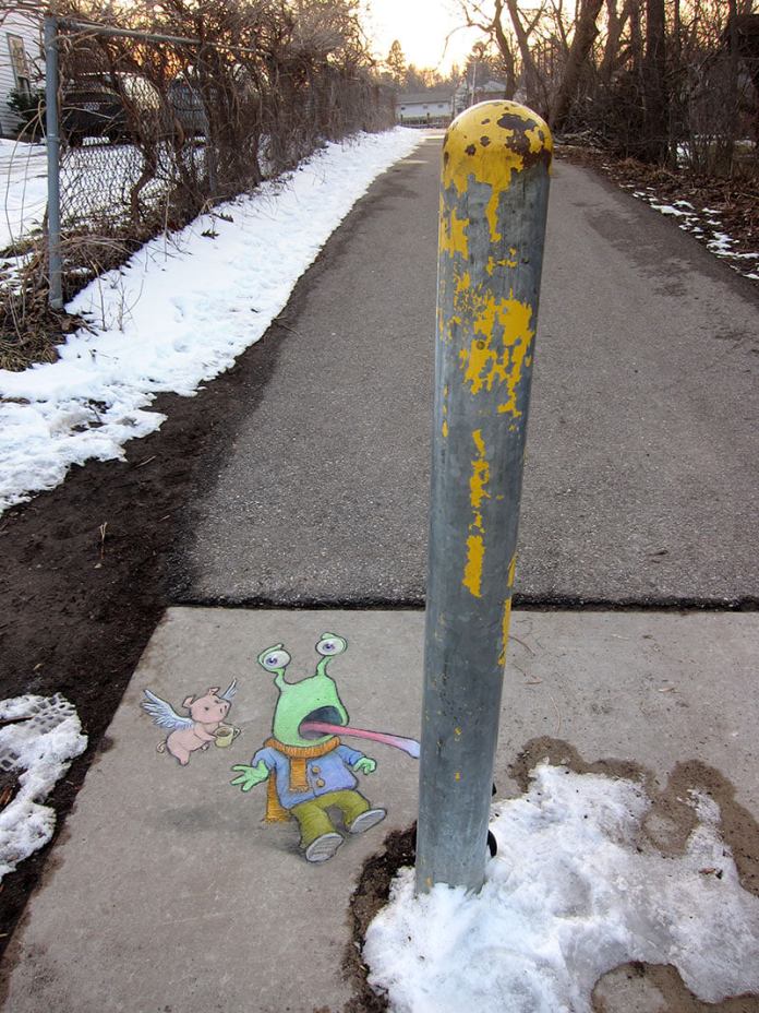 A whimsical chalk illustration by David Zinn titled "Be careful out there!" The artwork features a wide-eyed, green alien character with two stalked eyes and a long tongue, appearing startled and leaning back as if reacting to the yellow, weathered pole in front of it. The alien is dressed in a cozy outfit, including a scarf, jacket, and pants. Beside the alien is a flying pig holding a cup of coffee, adding an extra layer of humor and surreal charm. The scene is set on a snowy sidewalk path bordered by a chain-link fence and winter trees, with the setting sun casting a warm glow in the background.