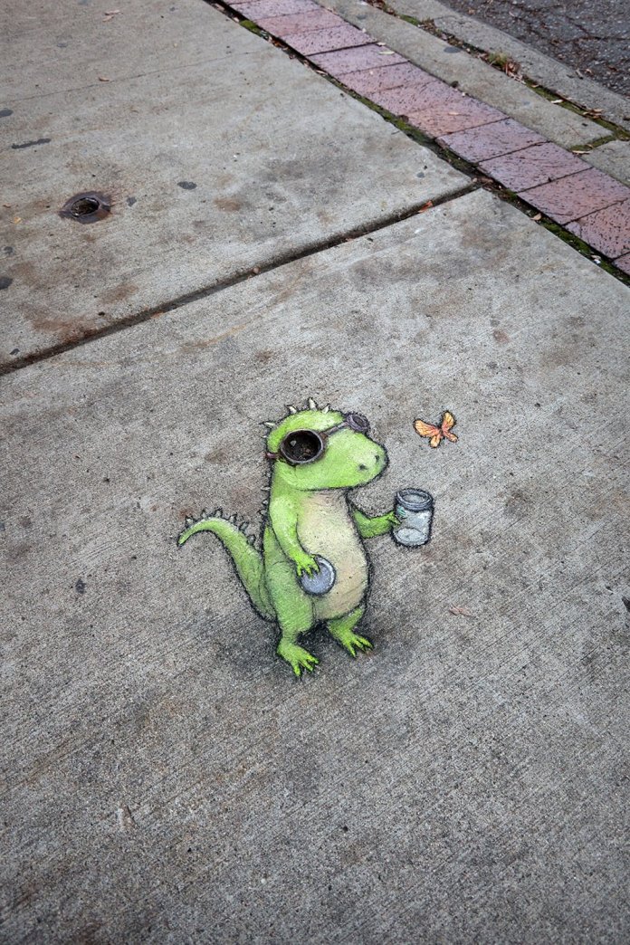 A charming chalk illustration by David Zinn titled "Thomas is studying the release and capture of small beautiful things." The artwork depicts a bright green lizard-like creature with textured skin and a curious expression. Thomas is wearing circular goggles, cleverly incorporating existing sidewalk features, and holding a jar in one hand as a delicate orange butterfly hovers nearby. The whimsical scene is drawn directly on the gray concrete sidewalk, with subtle shading adding depth to the lizard's body and the jar. A border of pink bricks and weathered pavement adds a natural urban frame to the artwork.