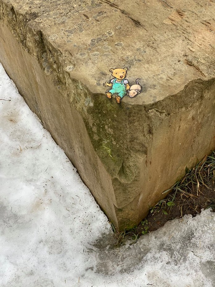 A delightful chalk drawing by David Zinn titled "Carl and Leslie waiting for the low ground to melt." The artwork is placed on the edge of a large, rough stone slab. Carl, a small teddy bear wearing teal overalls, sits comfortably, his expression calm and patient. Beside him is Leslie, a tiny, pink mouse with a curious demeanor. The drawing is perfectly integrated into the natural setting, with snow melting around the base of the stone, revealing patches of grass and earth. The contrast between the warm characters and the cold surroundings creates a whimsical and heartwarming scene.
