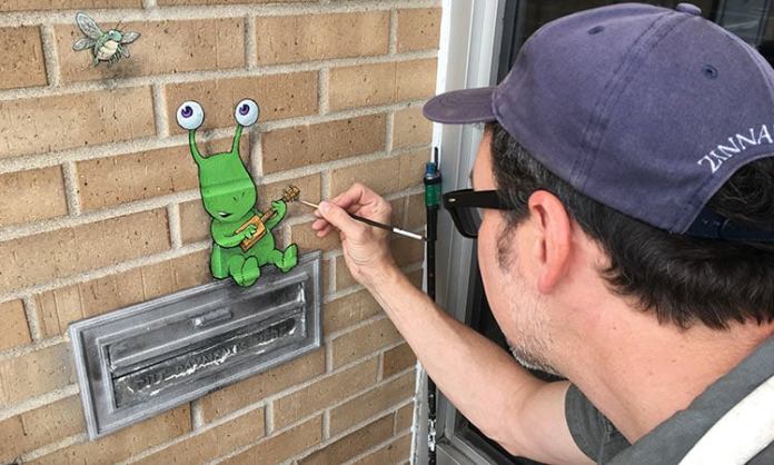 Artist David Zinn works on a whimsical chalk drawing on a brick wall. The artwork depicts a green alien with two antennae ending in expressive eyes, sitting atop a metal letterbox. The alien is strumming a small orange ukulele, adding a musical charm to the scene. A tiny fly with wings is drawn nearby on the wall, adding another playful element. Zinn, wearing a blue cap with the word "ZINNART" visible, carefully applies the finishing touches with a paintbrush. The warm tones of the brick wall serve as a perfect backdrop for the colorful, imaginative creation.