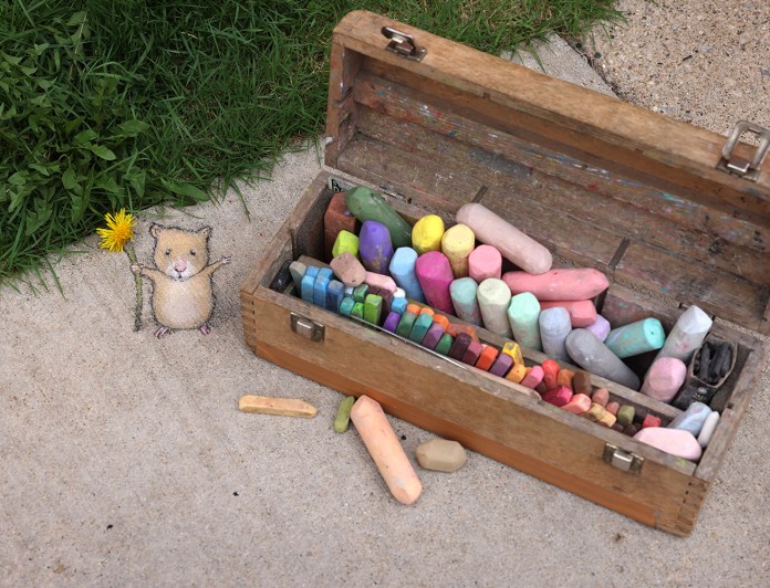 A whimsical chalk art scene by David Zinn featuring a tiny, cheerful creature holding a bright yellow dandelion, drawn on a concrete sidewalk. The character is playfully interacting with a wooden box filled with colorful chalk sticks in various sizes and hues, emphasizing the vibrant creativity of Zinn's work. The background includes patches of green grass, adding a natural element to the composition.