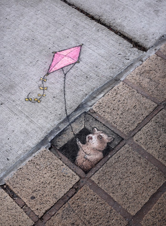 A charming chalk art piece by David Zinn featuring a small animal character peeking out from a square opening in the pavement, holding the string of a pink kite. The kite, drawn on the concrete sidewalk above, trails a string with yellow bows, creating a whimsical connection between the artwork and its surroundings. The scene integrates the textured bricks and cement of the environment, enhancing the illusion of depth and storytelling.