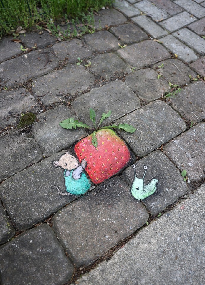 A delightful chalk art scene by David Zinn featuring a tiny mouse character in a teal outfit pushing a vibrant red strawberry, drawn on a cobblestone pathway. A real dandelion plant emerges naturally from the top of the strawberry, blending the artwork with its environment. Next to the mouse, a cute green snail with wide eyes adds a whimsical touch to the scene. The textured stones and surrounding greenery enhance the playful and imaginative quality of the composition.