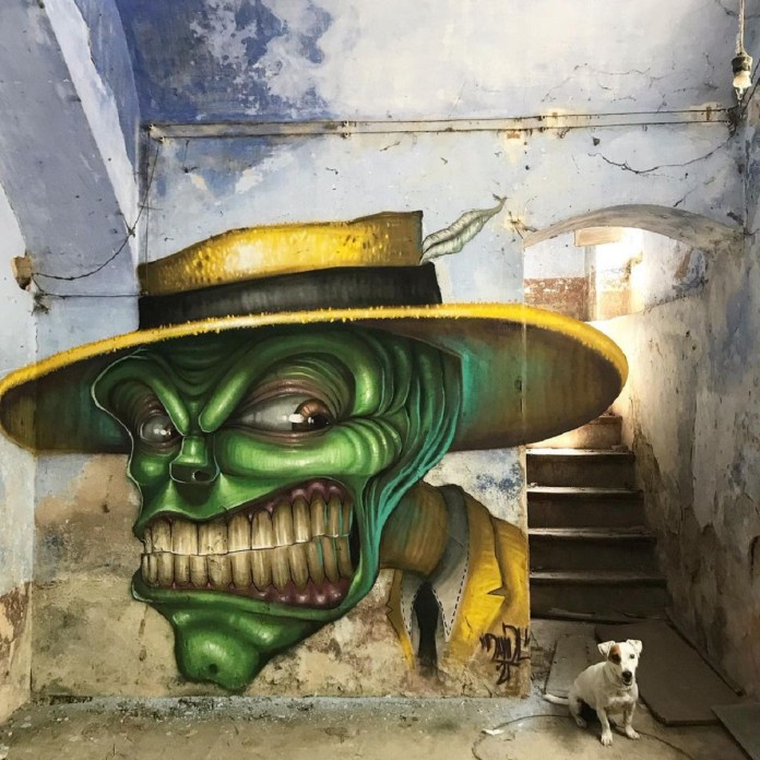 Post-graffiti artwork by DavidL featuring a green-faced, zombie-like character inspired by The Mask, wearing a yellow hat and suit, painted on a distressed wall with a small white dog sitting in front, located outside Barcelona, Spain.