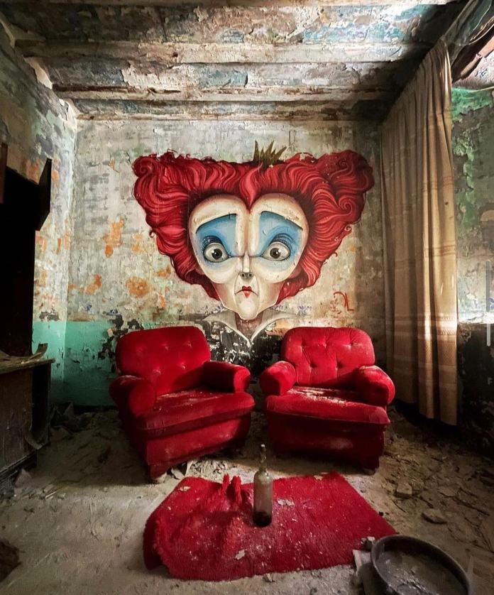 Post-graffiti artwork by DavidL featuring the Red Queen from Alice in Wonderland, painted on a distressed wall with two red armchairs and a bottle in the foreground, located outside Barcelona, Spain.